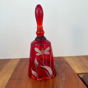 May include: A red glass bell with a dragonfly painted on the front. The bell has a scalloped edge and a clear glass handle.