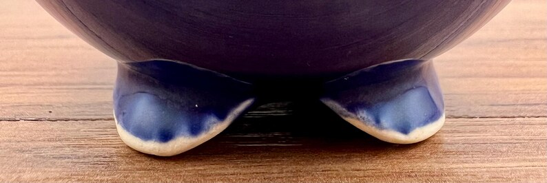 May include: A close-up of a dark blue ceramic bowl with two curved feet. The feet are a darker shade of blue with a white trim at the base. The bowl sits on a wooden surface.