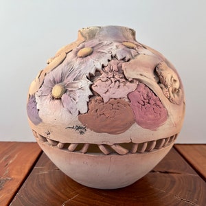 May include: A round ceramic vase with a textured surface and a floral design. The vase is decorated with pink, brown, and white flowers. The vase has a cut-out design around the bottom edge. The vase is signed "J. Davis 96".