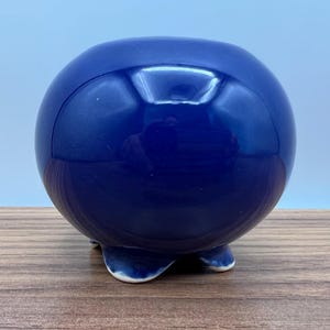 May include: A glossy, spherical, cobalt blue ceramic decorative object. The object has a rounded shape and rests on three small, curved feet. The surface reflects light, creating highlights. The object is placed on a wooden surface.