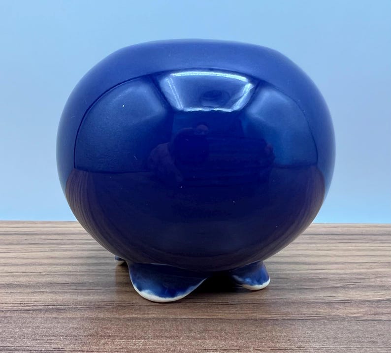 May include: A glossy, spherical, cobalt blue ceramic decorative object. The object has a rounded shape and rests on three small, curved feet. The surface reflects light, creating highlights. The object is placed on a wooden surface.