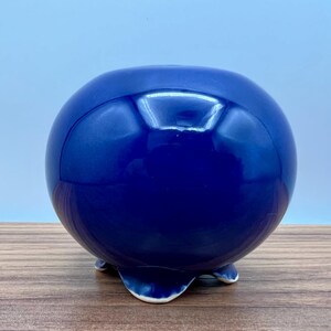May include: A glossy, spherical, cobalt blue ceramic planter. The round pot has a smooth surface and rests on three small, curved feet. The interior opening is visible at the top. The planter is suitable for small plants or succulents.