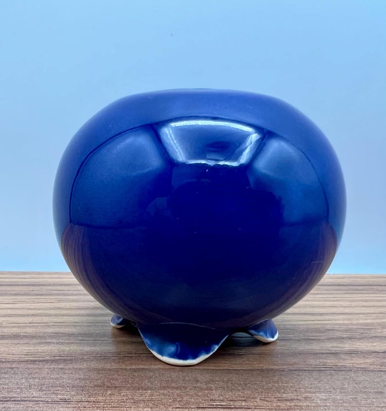 May include: A glossy, spherical, cobalt blue ceramic planter. The round pot has a smooth surface and rests on three small, curved feet. The interior opening is visible at the top. The planter is suitable for small plants or succulents.