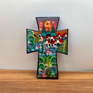 Peut inclure: Une croix décorative en bois avec une scène d'art populaire colorée. L'œuvre représente un village avec des maisons aux toits rouges, une église et des personnages portant des chapeaux. La croix a une bordure noire et est placée sur un fond neutre.