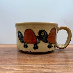 May include: A ceramic mug with a tan base colour and a brown handle. The mug features a repeating pattern of turtles in black, orange, and green. The mug is sitting on a wooden surface.