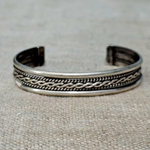 May include: A silver-toned cuff bracelet featuring a central twisted rope pattern flanked by smooth, polished bands. The open-ended design has a metallic finish with darkened recessed areas that highlight the intricate braided texture along the center.