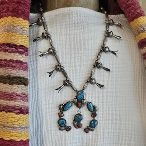 May include: A silver squash blossom necklace featuring a central naja pendant adorned with inlaid turquoise and coral stones. The necklace chain consists of round silver beads interspersed with silver blossom-shaped accents along the sides.