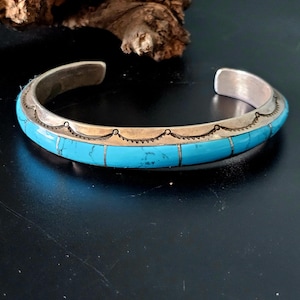 May include: Silver cuff bracelet with inlaid turquoise stones. The bracelet features a bright turquoise band with a silver border and decorative etching. The open-ended design allows for easy wear. A classic piece of Southwestern jewelry.