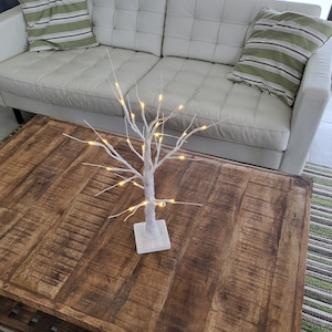 Puede incluir: Un árbol blanco en forma de rama con luces blancas cálidas sobre una mesa de café de madera. La mesa tiene un acabado rústico y desgastado.
