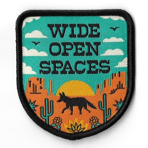 Puede incluir: Un parche bordado en negro y naranja con una escena de puesta de sol y el texto "WIDE OPEN SPACES". El parche presenta un coyote, cactus y una cordillera.