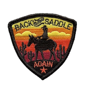 Pode incluir: Patch bordado com uma cena desértica com um cowboy a cavalo, cactos e um pôr do sol. O patch diz "BACK IN THE SADDLE AGAIN" em letras laranja e brancas, com uma borda preta.