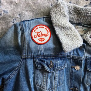Może przedstawiać: Niebieska kurtka dżinsowa z białym kołnierzem z imitacji futra. Kurtka ma czerwoną i białą naszywkę z napisem "Jolene" i czerwonym wzorem ust. Na kołnierzu znajduje się mała szpilka z tęczową g&oacute;rą.