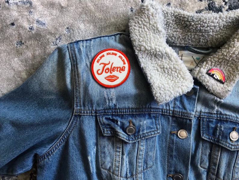 Może przedstawiać: Niebieska kurtka dżinsowa z białym kołnierzem z imitacji futra. Kurtka ma czerwoną i białą naszywkę z napisem "Jolene" i czerwonym wzorem ust. Na kołnierzu znajduje się mała szpilka z tęczową g&oacute;rą.