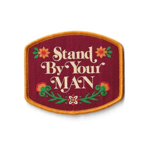 May include: An embroidered patch in burgundy with a gold border. The patch features a floral design and the text "Stand By Your MAN".