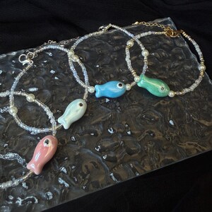 May include: Three beaded bracelets featuring small, glossy ceramic fish charms in pastel pink, light blue, and mint green. Each bracelet is constructed with translucent seed beads, small white faux pearls, and a gold-toned metal lobster clasp.