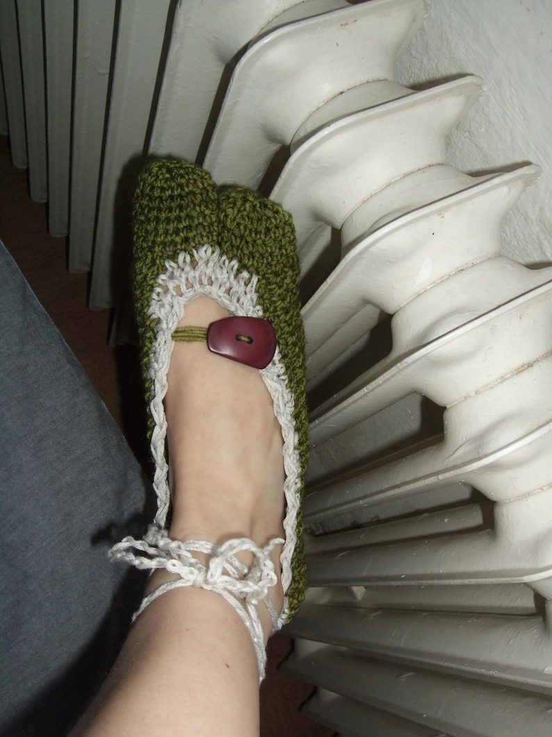 May include: Handmade green crochet slipper with white trim and a brown button closure. The slipper has a white tie closure around the ankle. The slipper is approximately 23 cm long.