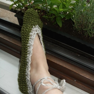 May include: A green and white crochet slipper with a lace trim and tie closure. The slipper is handmade and appears to be comfortable. The slipper is shown on a foot near a window with plants.