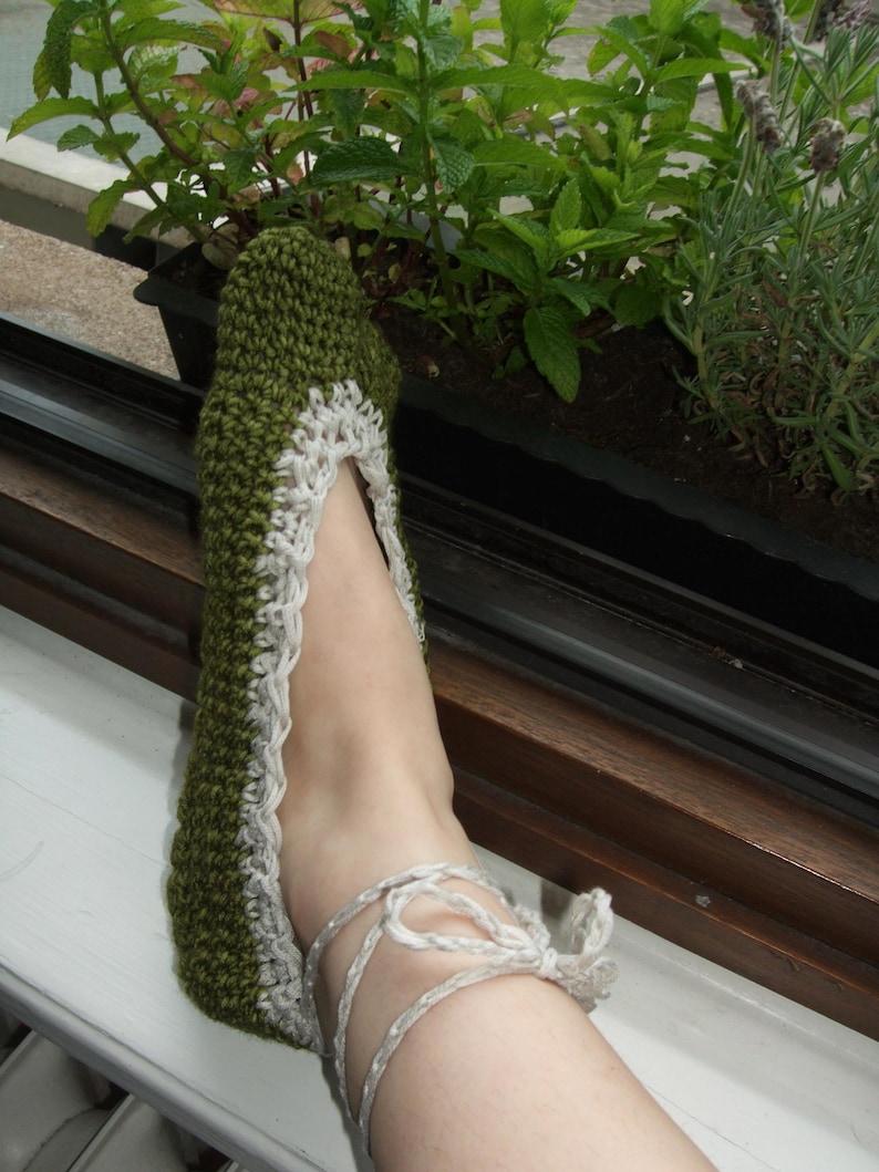 May include: A green and white crochet slipper with a lace trim and tie closure. The slipper is handmade and appears to be comfortable. The slipper is shown on a foot near a window with plants.