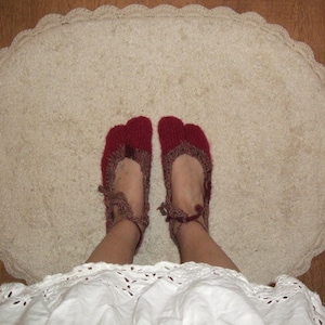 May include: Red and brown knitted slippers with a split toe, worn on a cream oval rug with a scalloped edge. The person is wearing a white dress. The slippers are a cozy footwear option.