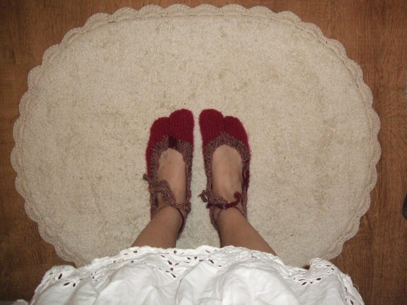 May include: Red and brown knitted slippers with a split toe, worn on a cream oval rug with a scalloped edge. The person is wearing a white dress. The slippers are a cozy footwear option.