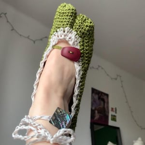 May include: Handmade green crochet sandals with a white trim and a maroon button closure. The sandals feature a unique design with a toe separator and a white crocheted ankle strap. A decorative iridescent square is attached to the ankle strap.