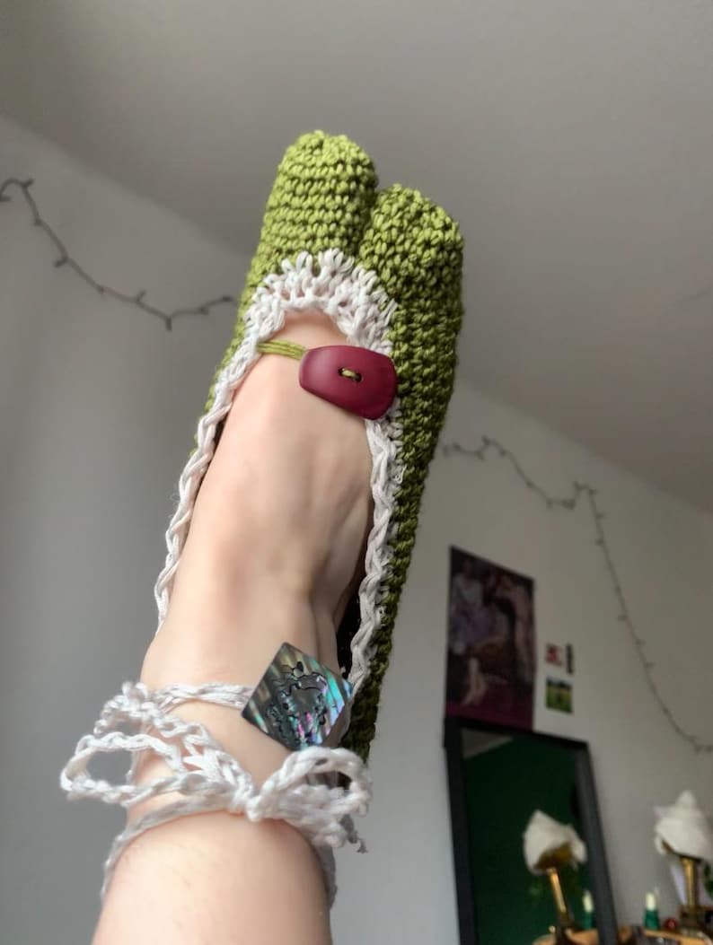 May include: Handmade green crochet sandals with a white trim and a maroon button closure. The sandals feature a unique design with a toe separator and a white crocheted ankle strap. A decorative iridescent square is attached to the ankle strap.