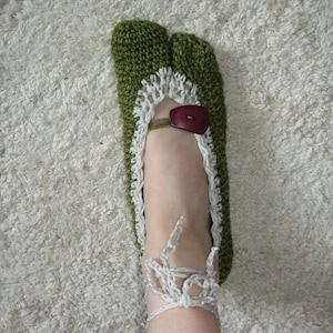 May include: Hand-knitted green and white slipper with a split toe design. The slipper features a white lace trim, a brown button closure, and a white ankle strap. The slipper is designed for indoor use.