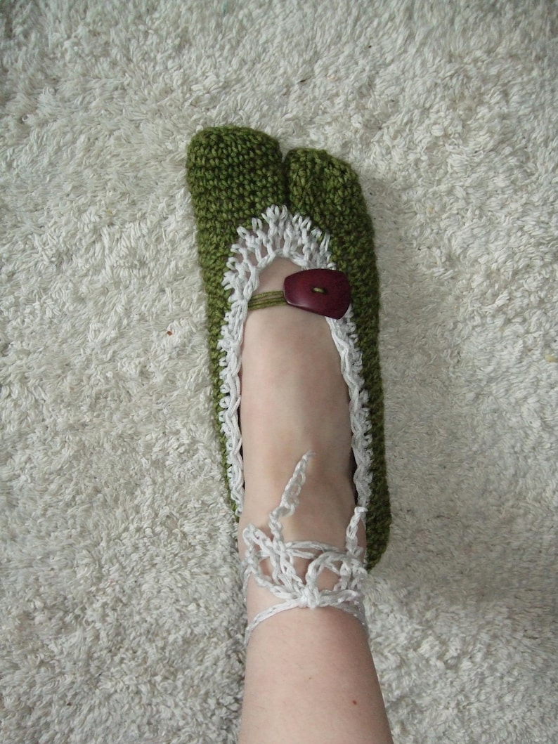 May include: Hand-knitted green and white slipper with a split toe design. The slipper features a white lace trim, a brown button closure, and a white ankle strap. The slipper is designed for indoor use.