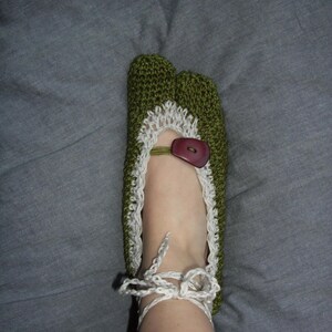 May include: Green crocheted slipper with white lace trim and a brown button. The slipper features a split toe design and white tie-up laces. The handmade slipper is designed to be worn indoors.