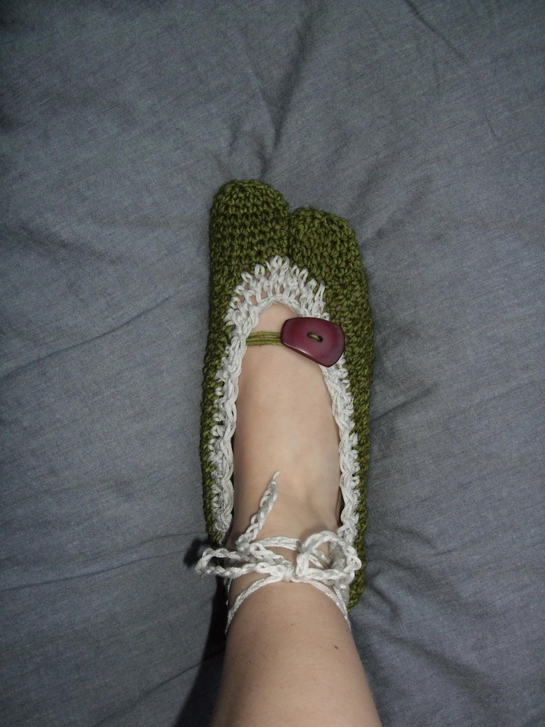 May include: Green crocheted slipper with white lace trim and a brown button. The slipper features a split toe design and white tie-up laces. The handmade slipper is designed to be worn indoors.