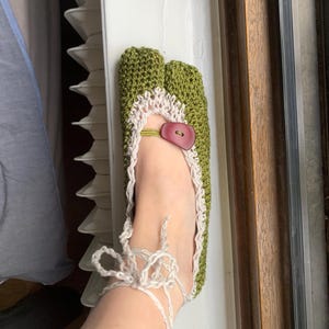 May include: Handmade green crochet slipper with a split toe design. The slipper features a cream-colored trim and a red button closure. White yarn ties secure the slipper around the ankle. A unique and cozy footwear item.
