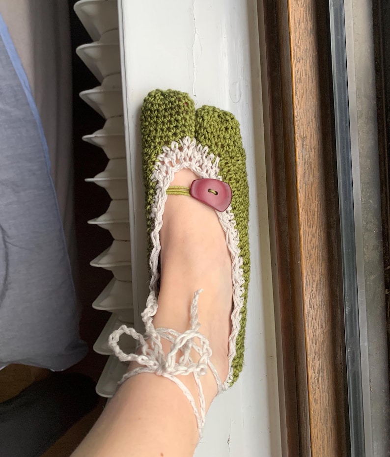 May include: Handmade green crochet slipper with a split toe design. The slipper has a cream-coloured trim and a red button closure. White yarn ties secure the slipper around the ankle. A unique and cosy footwear item.