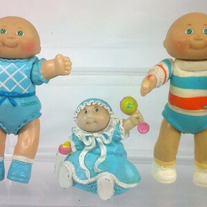 May include: Three vintage Cabbage Patch Kids dolls. One doll is holding a yellow spoon, another a red football, and the third a rattle. The dolls are wearing blue and white outfits. The dolls have bald heads, rosy cheeks, and green eyes.