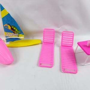 May include: A collection of Barbie beach-themed accessories in bright colors. Includes a pink boat, two pink lounge chairs, a small pink table with two white cups, and a sailboat with a toucan graphic.