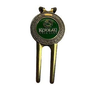 May include: A golf divot repair tool with a bronze finish. The tool features a circular emblem with the text "KO'OLAU GOLF CLUB" in white lettering on a green background. The emblem is surrounded by a decorative border.