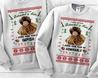 May include: Two gray Christmas sweaters with a red and white pattern. The sweaters feature a picture of a man dressed as a Christmas character with the text "Cheer or Fear Belsnickel is Here" and "I Judge Your Year As Impish" or "I Judge Your Year As Admirable".