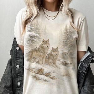 May include: Cream-colored t-shirt featuring a detailed illustration of two wolves in a snowy forest scene. The design includes evergreen trees, a river, and a winter landscape. The wolves are depicted with realistic fur and attentive expressions.