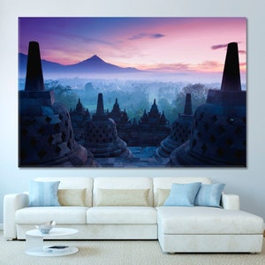 Op de afbeelding: Een panoramisch uitzicht op een boeddhistisch tempelcomplex bij zonsopgang. De tempel is gemaakt van steen en heeft veel ingewikkelde houtsnijwerk. De lucht is een prachtige mix van roze, paars en oranje. De tempel is omgeven door weelderige groene bomen en een mistige vallei.