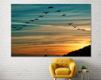 May include: A flock of geese flying in a V formation against a colorful sunset sky. The geese are silhouetted against the orange, yellow, and blue hues of the sky.