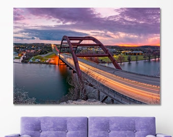 May include: A photograph of a red bridge with cars driving over it at sunset. The bridge is over a body of water and there are trees and buildings in the background.