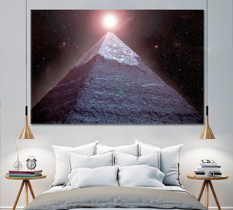 May include: A large canvas print of a pyramid under a starry night sky with a bright light at the top. The pyramid is a dark blue-gray color, and the sky is a deep purple with many stars. The print is displayed above a bed.
