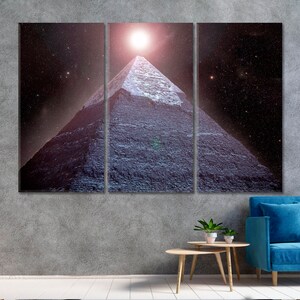 May include: A three-panel canvas print depicting a pyramid under a bright light in a starry night sky. The pyramid is constructed of dark gray stone blocks. The artwork is displayed above a blue sofa and a small wooden table.