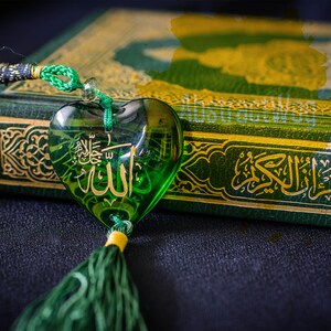 May include: A green heart-shaped charm with the word "Allah" inscribed in Arabic script hangs from a green cord. The charm is resting on a green book with gold lettering and intricate designs. The book is titled "Al Quran".