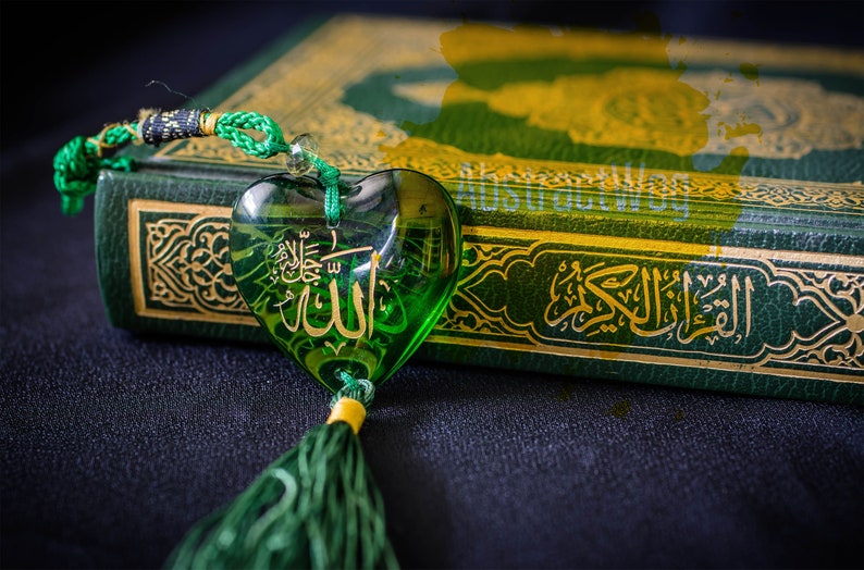 May include: A green heart-shaped charm with the word "Allah" inscribed in Arabic script hangs from a green cord. The charm is resting on a green book with gold lettering and intricate designs. The book is titled "Al Quran".