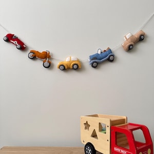 Puede incluir: Una guirnalda de vehículos de juguete de fieltro en rojo, naranja, amarillo, azul y beige cuelga sobre un camión de juguete de madera. La guirnalda incluye un coche de carreras, una moto, un coche, un camión y otro coche.
