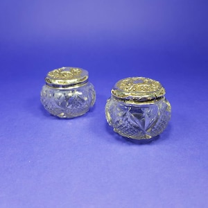 Pode incluir: Dois pequenos frascos de vidro transparente com tampas douradas ornamentadas. Os frascos têm um design de vidro lapidado e as tampas apresentam padrões intrincados. Os frascos estão sobre um fundo azul sólido, sugerindo uma estética vintage ou antiga.