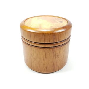 May include: A round wooden box with a lid. The box is made of light brown wood with a darker brown wood trim around the top and bottom. The box is approximately 10 cm in diameter and 7.5 cm tall.