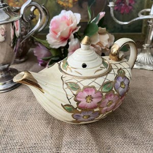 May include: A white teapot with a gold trim and floral design. The teapot has a lid with a small hole in the top. The teapot is sitting on a brown hessian surface.