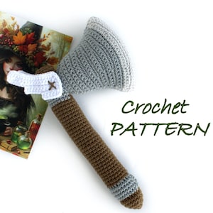 Puede incluir: Patrón de crochet para un hacha gris y marrón. El hacha está hecha de hilo y tiene un mango blanco con una cruz marrón.