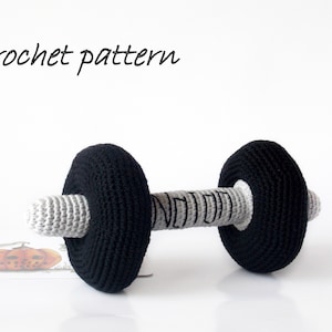 May include: Crochet pattern for a gray and black dumbbell. The dumbbell is made of yarn and has a black and white striped handle.